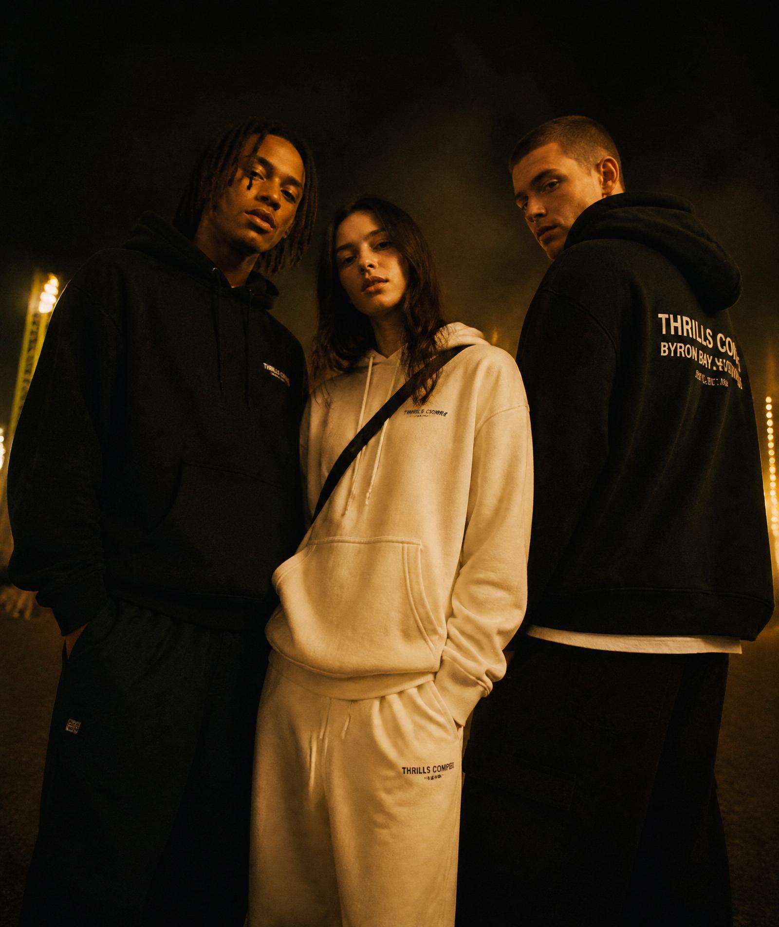 Three young adults in oversized black and cream streetwear under night festival lights